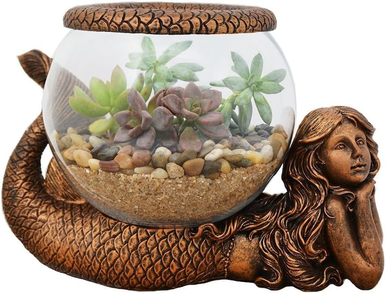 May include: A bronze-colored mermaid figurine cradles a clear glass terrarium filled with succulents, sand, and small pebbles. The mermaid has long flowing hair and is resting her head on her hands. The terrarium is round with a decorative rim.