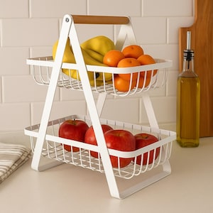 May include: A two-tiered white wire fruit basket with a wooden handle. The top basket holds bananas and oranges, while the bottom basket contains red apples. The basket is designed for kitchen or dining room use.