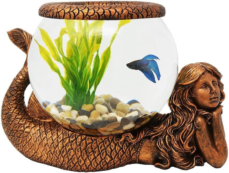 May include: A bronze-colored mermaid-shaped aquarium. The fishbowl is held by the mermaid's tail and arms. Inside the bowl are small pebbles, green plants, and a blue betta fish. The top of the bowl has a decorative, textured rim.