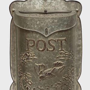 May include: A galvanized metal mailbox with a decorative bird and floral design. The word "POST" is embossed on the front. The mailbox has a hinged lid and a keyhole for security. The overall shape is rectangular with a curved top.