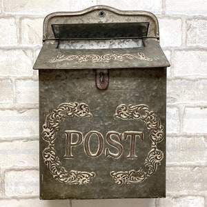 May include: A wall-mounted galvanized metal mailbox with a weathered gray finish. The front features the word POST in raised capital letters, surrounded by an embossed scrollwork frame. It includes a hinged top lid and a central latch.