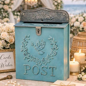 May include: A light blue metal mailbox with a decorative dove and wreath design. The word "POST" is embossed on the front. The mailbox has a hinged lid and a lock. The background includes flowers, candles, and a sign that says "Cards & Wishes".