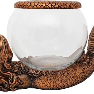 May include: A decorative glass bowl supported by a bronze-colored mermaid sculpture. The mermaid is lying down, with her tail curved to cradle the bowl. The bowl has a textured lid.