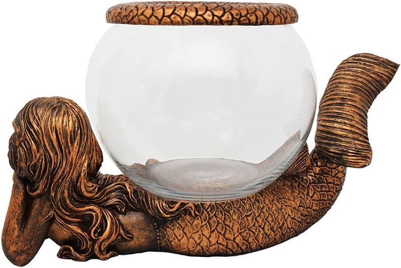 May include: A decorative glass bowl supported by a bronze-colored mermaid sculpture. The mermaid is lying down, with her tail curved to cradle the bowl. The bowl has a textured lid.