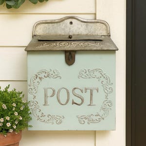 May include: A light blue metal mailbox with a hinged lid and the word "POST" in large letters. The mailbox has decorative scrollwork and is mounted on a white wall. A small potted plant sits to the left.