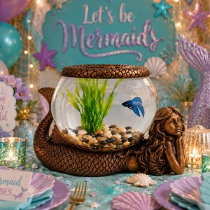 May include: A mermaid-themed centerpiece features a fishbowl with a blue betta fish, resting on a bronze mermaid sculpture. The scene includes a sign that says "Let's be Mermaids", seashells, and teal and purple accents, creating a whimsical underwater vibe.