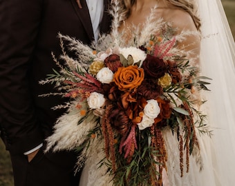May include: A wedding couple standing together. The individual on the left wears a dark brown suit with a white shirt, beige bowtie, and a floral boutonniere. The individual on the right wears a white lace wedding dress and a long veil.
