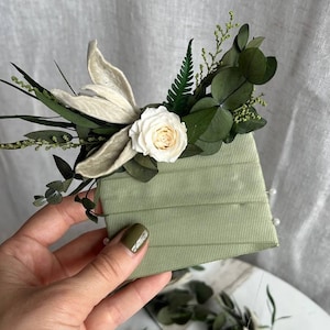 Könnte beinhalten: Eine salbeigrüne, rechteckige Bandkarte, verziert mit einem dekorativen Arrangement aus getrockneten weißen Rosen, grünen Eukalyptusblättern, Farnwedeln und beigem Blattwerk. Das Band ist horizontal um die Karte gewickelt und mit Nadeln fixiert.