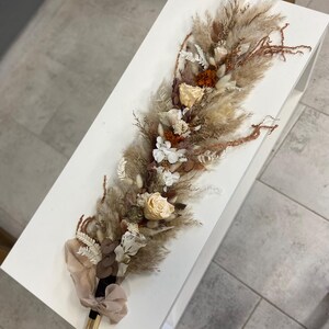May include: A long, narrow floral arrangement featuring dried pampas grass, beige and white roses, and rust-colored accents. The stems are gathered at the base and tied with a light beige ribbon, creating a textured, elongated bouquet of dried plants.