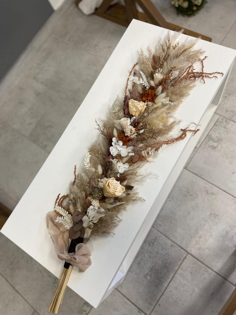 May include: A long, narrow floral arrangement featuring dried pampas grass, beige and white roses, and rust-colored accents. The stems are gathered at the base and tied with a light beige ribbon, creating a textured, elongated bouquet of dried plants.
