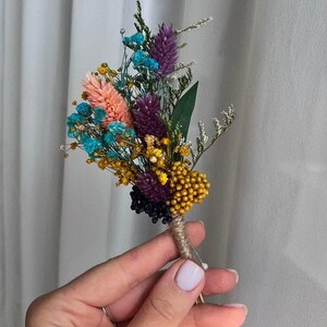 May include: A boutonniere featuring a collection of dried flowers including purple gomphrena, bright turquoise baby's breath, a peach-colored plume, yellow yarrow, and green foliage. The stems are wrapped in natural tan twine with a silver pin.