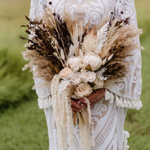 May include: A dried floral bouquet featuring cream-colored roses, white protea flowers, fluffy pampas grass, and dark brown foliage. The arrangement includes long, cascading white amaranth stems and is held against a white lace-patterned dress.