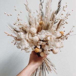 A beautiful handmade bridal bouquet featuring natural dried wildflowers in soft beige tones. This rustic bouquet is perfect for a wedding, adding a touch of nature to your special day. Ideal for the bride and bridesmaids, it captures the essence of earthy elegance with its delicate, dried floral arrangement. A charming and unique choice for those seeking a timeless and eco-friendly floral decor for their wedding day.