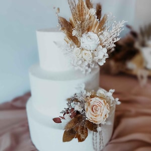 Op de afbeelding: Een witte taart met drie lagen, versierd met gedroogde bloemstukken. De bovenste laag bevat een boeket van witte rozen, pampasgras en palmbladeren. De middelste laag is versierd met beige rozen en gedroogd bladgroen.