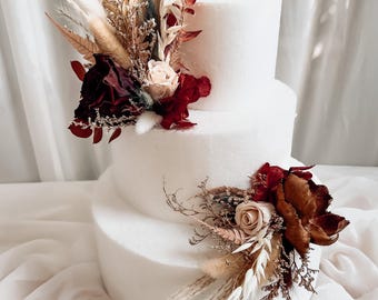 May include: A two-tier white cake decorated with dried floral arrangements. The top tier features a cluster of burgundy, cream, and tan dried flowers and grasses. The bottom tier has a smaller matching arrangement of dried roses and foliage.