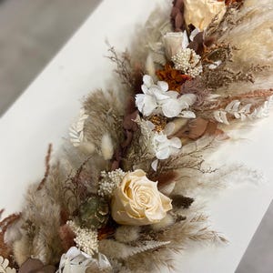 May include: A long floral arrangement featuring preserved cream roses, white hydrangea blossoms, fluffy pampas grass, and dried foliage in shades of brown, tan, and beige. The composition includes various textures of dried grasses and small white filler flowers.
