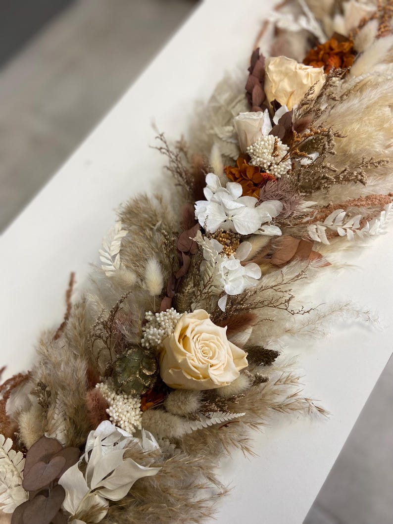May include: A long floral arrangement featuring preserved cream roses, white hydrangea blossoms, fluffy pampas grass, and dried foliage in shades of brown, tan, and beige. The composition includes various textures of dried grasses and small white filler flowers.