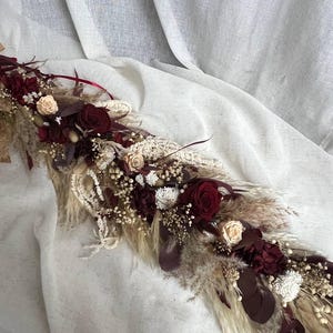 May include: A floral arrangement featuring deep burgundy roses, cream-colored roses, white dried flowers, and beige pampas grass. The composition includes dark red foliage and delicate filler flowers, creating a long, textured decorative piece.