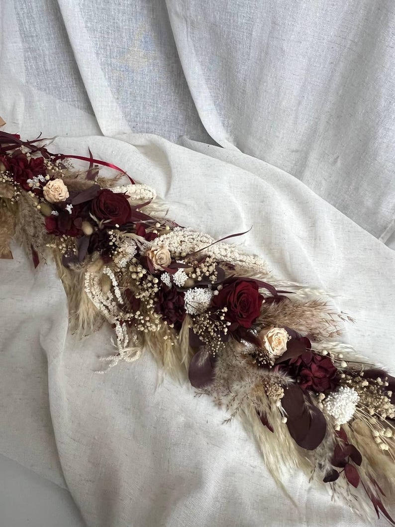 May include: A floral arrangement featuring deep burgundy roses, cream-colored roses, white dried flowers, and beige pampas grass. The composition includes dark red foliage and delicate filler flowers, creating a long, textured decorative piece.