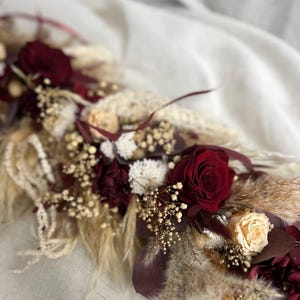 May include: A floral arrangement featuring deep burgundy roses, cream-colored roses, and dried pampas grass. The composition includes white baby's breath, dark maroon foliage, and various dried botanical elements arranged in a long, linear display.