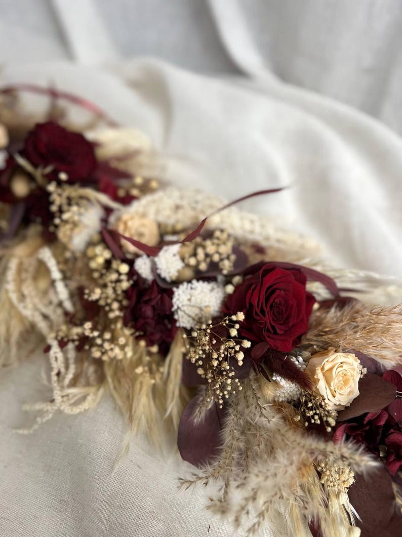 May include: A floral arrangement featuring deep burgundy roses, cream-colored roses, and dried pampas grass. The composition includes white baby's breath, dark maroon foliage, and various dried botanical elements arranged in a long, linear display.