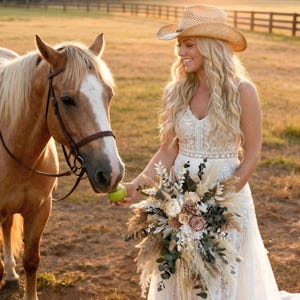 May include: A light-colored horse with a brown leather bridle and a person wearing a straw cowboy hat and a white lace dress. The person holds a green apple for the horse and carries a large bouquet of dried flowers, pampas grass, and greenery.