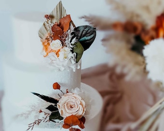 May include: A multi-tiered white cake decorated with dried and preserved floral arrangements. The top tier features a fan-shaped palm leaf, white roses, and rust-colored foliage. The lower tier includes a large pale peach rose and dark green leaves.