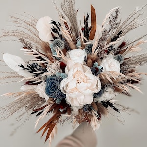Op de afbeelding: Een bloemenboeket met grote witte pioenrozen, een enkele stoffig blauwe roos en diverse gedroogde elementen, waaronder beige pampasgras, bruine palmbladeren, donker zwart blad en kleine blauwe distelkoppen in een dichte structuur.