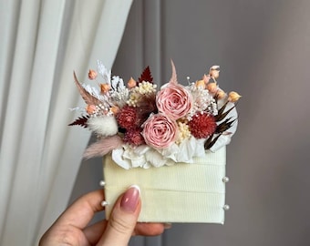 May include: A rectangular cream-colored box with a ribbed texture, adorned with small white pearls on the sides. The top is decorated with an arrangement of dried flowers, including two pink roses, red globe amaranth, white baby's breath, and grasses.