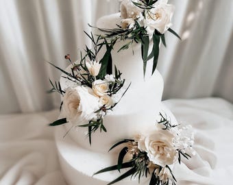 May include: A three-tiered white cake decorated with arrangements of cream-coloured roses, small white flowers, and dark green foliage. The floral clusters are placed asymmetrically on each tier, creating a cascading effect down the side of the cake.