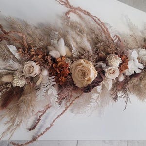 May include: A floral arrangement featuring dried pampas grass, preserved roses in shades of cream and beige, white ferns, and brown foliage. The elongated display includes soft textures and a long, light beige ribbon attached to one end.