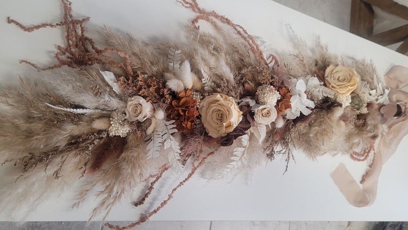 May include: A floral arrangement featuring dried pampas grass, preserved roses in shades of cream and beige, white ferns, and brown foliage. The elongated display includes soft textures and a long, light beige ribbon attached to one end.