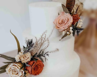 May include: A three-tiered white cake decorated with arrangements of dried flowers and foliage. The floral clusters include muted pink, rust orange, and cream roses, small white filler flowers, dark green leaves, and a single brown patterned feather.