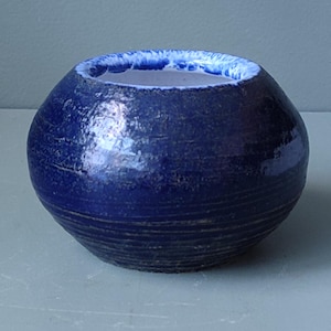 Puede incluir: Un cuenco pequeño y redondo de cerámica, de color azul oscuro brillante. La superficie texturizada presenta líneas horizontales. El borde interior es de un azul más claro. La forma general es esférica con una amplia abertura.