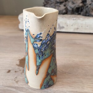 Puede incluir: Una jarra de cerámica con base blanca y un diseño abstracto en azul, verde y marrón.