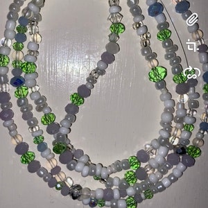 Puede incluir: Un collar de cuentas de múltiples hilos con cuentas blancas, verdes y moradas. El collar está hecho de cuentas de varios tamaños y formas.