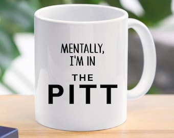 May include: White ceramic coffee mug with the text "MENTALLY, I'M IN THE PITT" in black, centered on the mug. The mug has a curved handle and is set against a blurred green background.