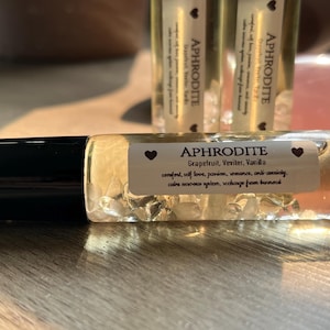 May include: A clear glass roller bottle with a black cap and a white label that reads "APHRODITE Grapefruit, Vetiver, Vanilla comfort, self love, passion, romance, anti-anxiety, calms nervous system, recharge from burnout".