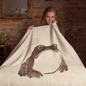 May include: A white fleece blanket with a brown frog illustration and a red stamp in the upper left corner.