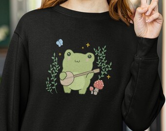 Könnte beinhalten: Schwarzes Sweatshirt mit einem grünen Frosch, der in einer Waldkulisse Banjo spielt. Der Frosch ist umgeben von grünen Blättern, einem blauen Schmetterling und roten Pilzen.
