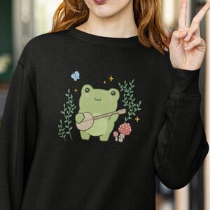 May include: Black sweatshirt with a green frog playing a banjo in a forest setting. The frog is surrounded by green leaves, a blue butterfly, and red mushrooms.