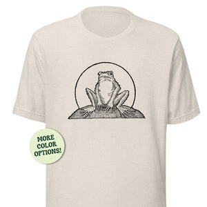 May include: A beige t-shirt with a black and white illustration of a frog sitting on a rock. The frog is facing forward with its mouth closed. The text "MORE COLOR OPTIONS!" is printed on a green circle in the lower left corner of the image.