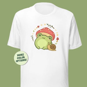 Könnte beinhalten: Weißes T-Shirt mit einem grünen Frosch, der einen roten und weißen Pilzhut trägt. Der Frosch sitzt neben einer braunen Schnecke. Das Design ist von grünen Blättern und weißen Blumen umgeben.