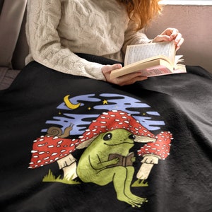 Peut inclure: Une couverture en polaire noire avec une illustration fantaisiste d'une grenouille verte assise sous un champignon rouge et blanc, en train de lire un livre. La grenouille est entourée d'autres champignons et d'un escargot. L'arrière-plan est un ciel bleu foncé avec une lune croissante et des étoiles.