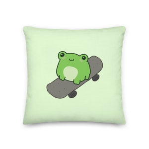 Könnte beinhalten: Ein grüner Frosch mit einem süßen Ausdruck sitzt auf einem grauen Skateboard mit schwarzen Rädern. Der Frosch befindet sich auf einem hellgrünen Hintergrund.