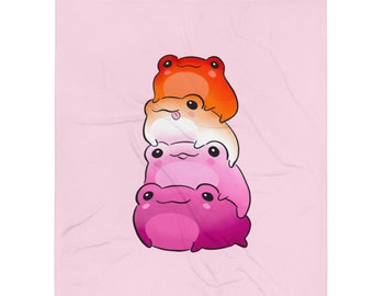 Op de afbeelding: Een roze deken met een ontwerp van drie cartoonkikkers die op elkaar zijn gestapeld. De kikkers zijn in tinten oranje, wit en roze. De deken heeft een zacht, getextureerd uiterlijk en staat tegen een lichtroze achtergrond.