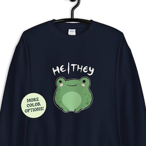 May include: Navy blue sweatshirt featuring a green frog and the text "He/They" in white. A green circle with the text "More Color Options!" is in the bottom left corner.