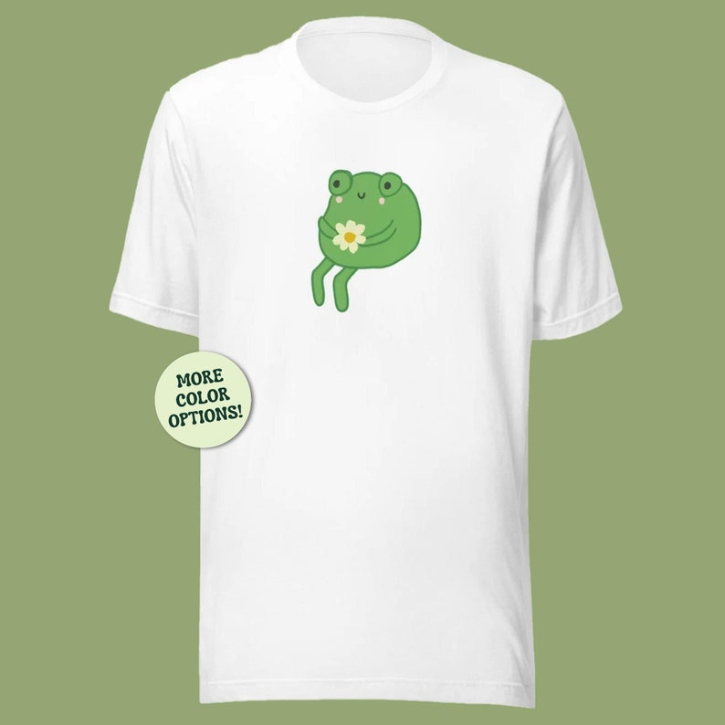 May include: White t-shirt with a green frog holding a white flower. The frog has a happy expression and is sitting on its hind legs. The text "MORE COLOR OPTIONS!" is on a green circle in the lower left corner of the image.