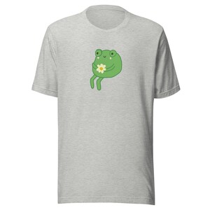 May include: A gray t-shirt with a green frog holding a white flower. The frog has a happy expression and is sitting on its hind legs.