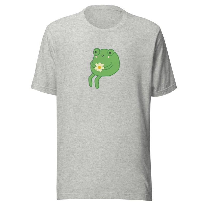 May include: A gray t-shirt with a green frog holding a white flower. The frog has a happy expression and is sitting on its hind legs.